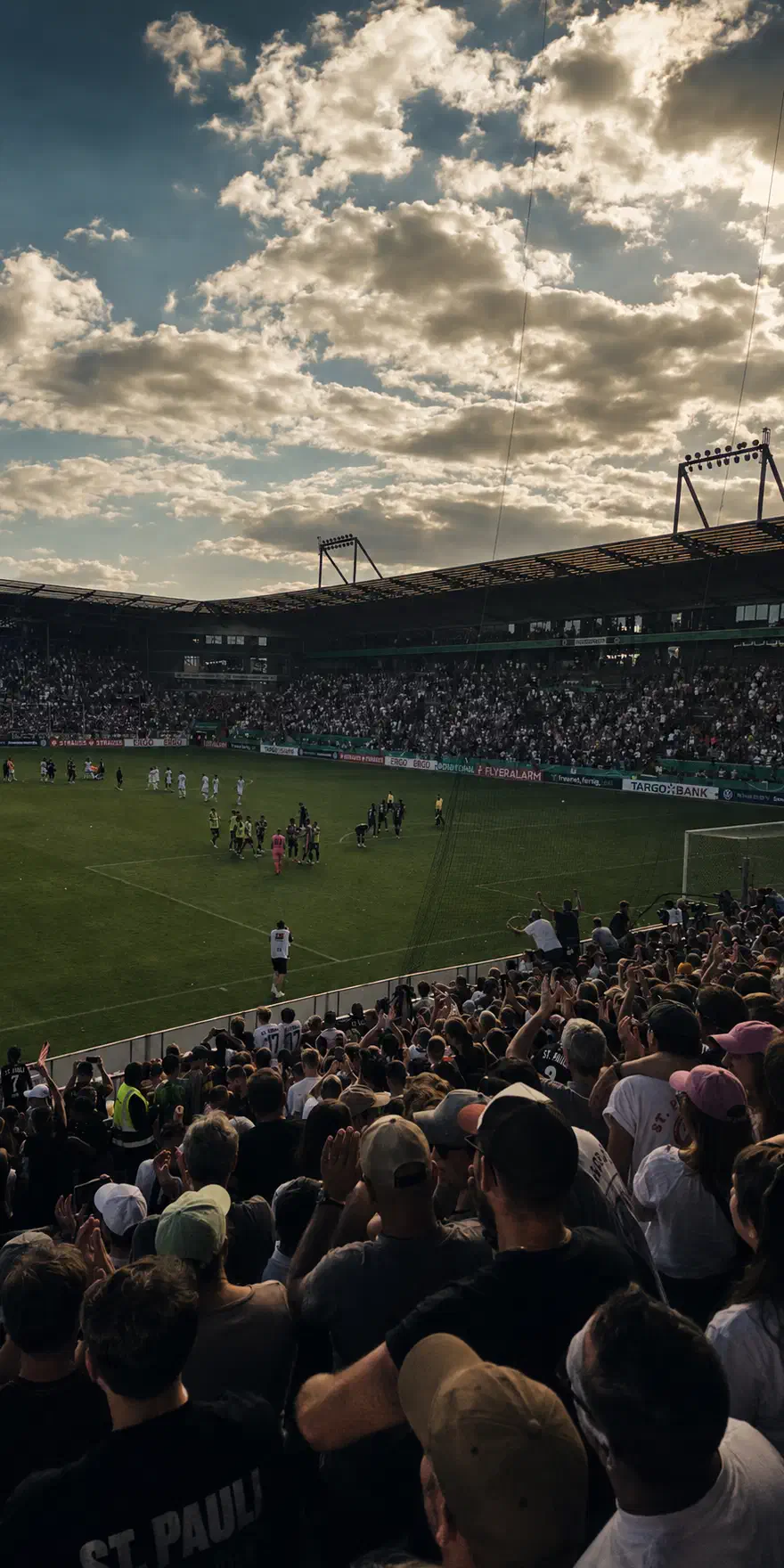 Volles Millerntor-Stadion im Abendlicht: Fankurve im Vordergrund, Mannschaft auf dem Rasen